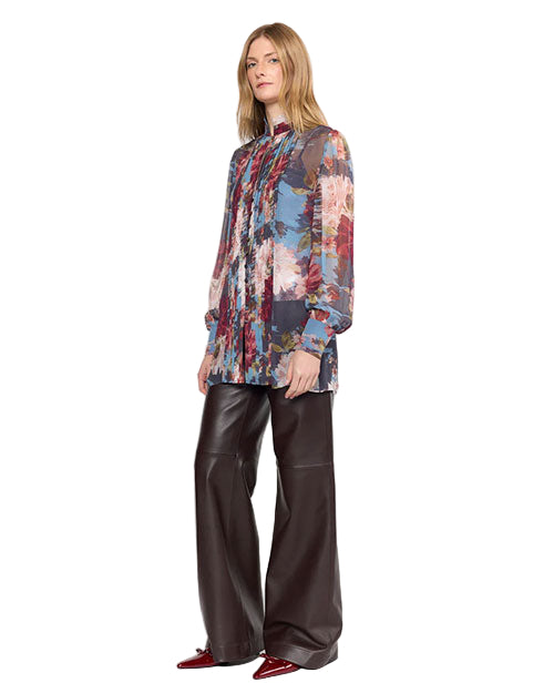A person stands wearing a stylish outfit featuring a long-sleeved, sheer blouse with a colorful abstract floral pattern in shades of blue, red, pink, and white. The blouse has a pleated front and a high neckline. They pair it with wide-leg, high-waisted black leather pants and shiny burgundy pointed-toe shoes.