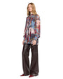 A person stands wearing a stylish outfit featuring a long-sleeved, sheer blouse with a colorful abstract floral pattern in shades of blue, red, pink, and white. The blouse has a pleated front and a high neckline. They pair it with wide-leg, high-waisted black leather pants and shiny burgundy pointed-toe shoes.
