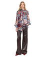 A person wearing a sheer, long-sleeved blouse with a vibrant abstract floral pattern in red, blue, pink, and white. The blouse has a high neckline and is paired with high-waisted, wide-leg black leather pants. They complete the look with pointed-toe, glossy red shoes. 