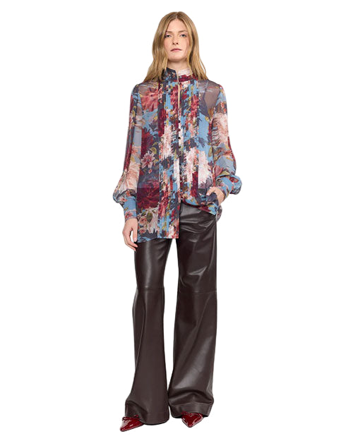 A person wearing a sheer, long-sleeved blouse with a vibrant abstract floral pattern in red, blue, pink, and white. The blouse has a high neckline and is paired with high-waisted, wide-leg black leather pants. They complete the look with pointed-toe, glossy red shoes. 