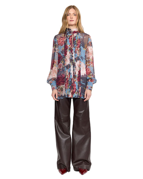 A person stands wearing a fashionable outfit that includes a sheer, long-sleeved blouse with a high neckline and a vibrant floral pattern in red, blue, pink, and green. The blouse features vertical pleats down the front. They pair it with high-waisted, wide-leg dark brown leather pants that have a voluminous silhouette, and glossy burgundy pointed-toe shoes.