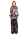 A person stands wearing a fashionable outfit that includes a sheer, long-sleeved blouse with a high neckline and a vibrant floral pattern in red, blue, pink, and green. The blouse features vertical pleats down the front. They pair it with high-waisted, wide-leg dark brown leather pants that have a voluminous silhouette, and glossy burgundy pointed-toe shoes.
