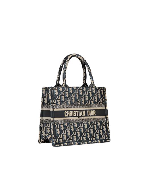 Side view of a Christian Dior designer tote bag. The bag features a repeating pattern of the brand’s initials, ‘CD,’ in a gray and black color scheme. A black stripe runs across the middle with the full brand name ‘CHRISTIAN DIOR’ printed in white capital letters. The bag has two black handles attached at the top for carrying.