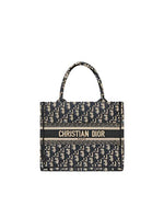 A Christian Dior designer tote bag. The bag features a repeating pattern of the brand’s initials, ‘CD,’ in a gray and blue ecru color scheme. A blue ecru stripe runs across the middle with the full brand name ‘CHRISTIAN DIOR’ printed in white capital letters. The bag has two black handles attached at the top for carrying.