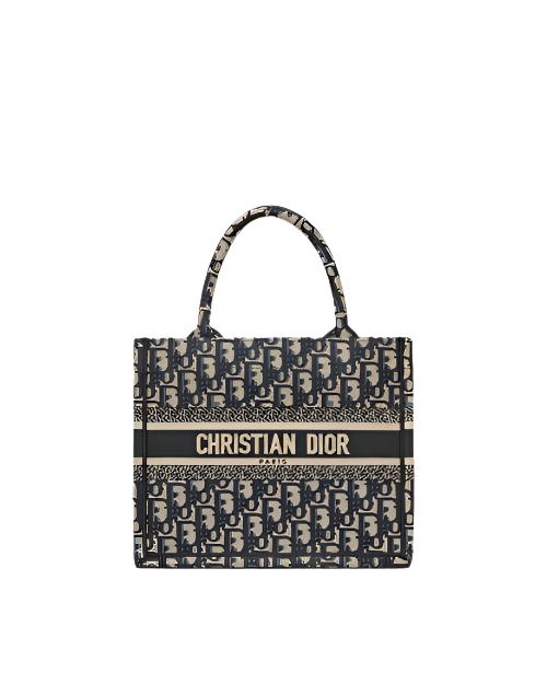 A Christian Dior designer tote bag. The bag features a repeating pattern of the brand’s initials, ‘CD,’ in a gray and blue ecru color scheme. A blue ecru stripe runs across the middle with the full brand name ‘CHRISTIAN DIOR’ printed in white capital letters. The bag has two black handles attached at the top for carrying.