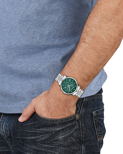 Person wearing a silver watch with a green face on a blue shirt and jeans background