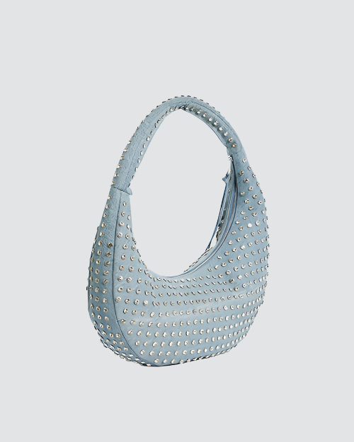 A side view of a large denim, crescent-shaped shoulder handbag with a single strap, adorned with numerous small, silver-colored studs creating a dotted pattern across the entire surface. 