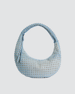 A large denim, crescent-shaped shoulder handbag with a single strap, adorned with numerous small, silver-colored studs creating a dotted pattern across the entire surface. 