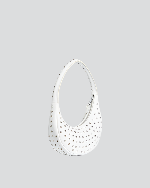 A side view of a white, crescent-shaped handbag with a single shoulder strap, adorned with numerous small, round silver studs. The bag features a zipper closure with tassel details on the side. 