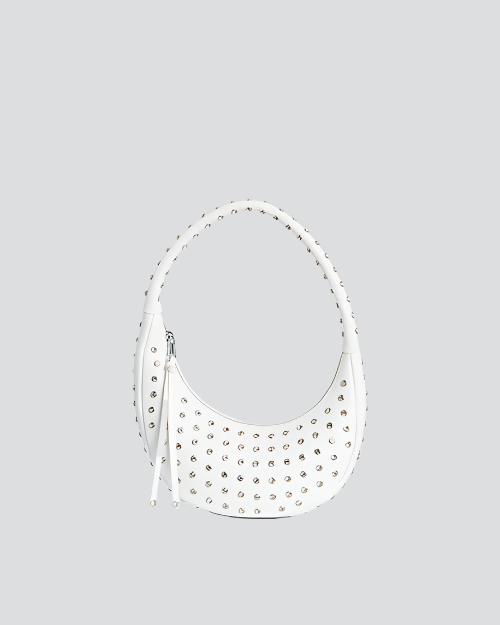 A white, crescent-shaped handbag with a single shoulder strap, adorned with numerous small, round silver studs. The bag features a zipper closure with tassel details on the side. 