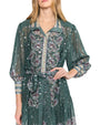 Close up of a person wearing a green long-sleeved dress with a paisley pattern in purple, white, and gold. The dress features a collar, button-down front, a belt tied at the waist, and slightly sheer sleeves with matching patterned cuffs.