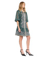 A person standing and wearing a green dress with intricate floral patterns and embellishments. The dress features three-quarter length sleeves, a collar, and buttons down the front. The person is also wearing black high-heeled shoes with ankle straps. The image is taken from the front-right angle, showcasing the dress's design and details.