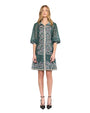 A person wearing a green dress with intricate floral embroidery and embellishments. The dress features sheer elbow-length sleeves, a collar, and buttons down the front, with a hemline that falls above the knees. The person is also wearing black pointed-toe heels with ankle straps.