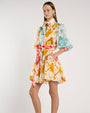 Person standing in a studio against a light neutral background, wearing a short, collared shirt dress with elbow-length sleeves and a tied waist sash. The dress features an abstract print in teal, mustard yellow, coral, and white. 