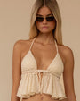 A model wearing a cream-colored textured top with a halter neckline that ties at the back of the neck. The top features two triangular cups made from a nubby, boucle-like fabric. Below the bust, the material flares into a loose, ruffled peplum with soft, wavy folds. A small tie at the center front connects the cups and adds an adjustable detail.