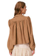Back view of a person wearing a light brown long-sleeved jacket with gathered fabric details at the shoulders and upper back, paired with a white lace skirt. 