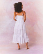Back view of a white, mid-calf length dress with thin straps, lace and embroidery. The dress has a fitted bodice and flows out into a fuller skirt at the bottom.  