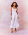 A model in a white, mid-calf button down length dress with thin straps, lace and embroidery. The dress has a fitted bodice and flows out into a fuller skirt at the bottom. 
