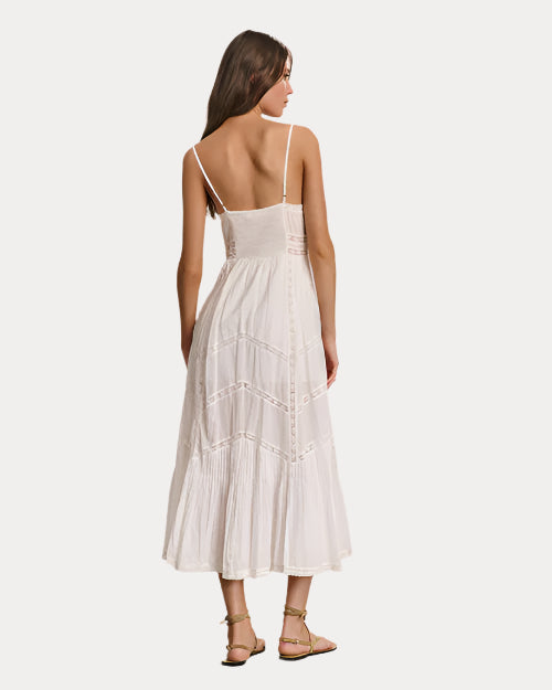A backside view of a brunette model with her hair pushed back wearing a white, sleeveless, ankle-length dress with thin shoulder straps, and a low neckline. The dress has a flowy skirt with zig-zag lace detailing. The model is also wearing beige strapped sandals and standing against a white background.