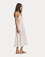 A sideview of a brunette model wearing a white, sleeveless, ankle-length dress with thin shoulder straps, V-neckline, and linear detailing at the waist. The dress has buttons running down the front and a flowy skirt with zig-zag lace detailing. The model is also wearing beige strapped sandals and standing against a white background.