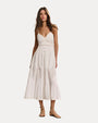 A brunette model wearing a white, sleeveless, ankle-length dress with thin shoulder straps, V-neckline, and linear detailing at the waist. The dress has buttons running down the front and a flowy skirt with zig-zag lace detailing. The model is also wearing beige strapped sandals and standing against a white background.