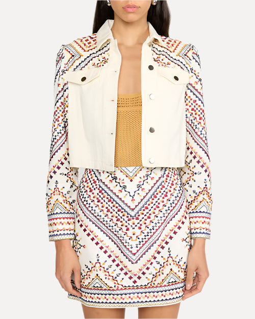 A body shot of a person wearing a yellow shirt under a white denim jacket with intricate, colorful embroidery on the shoulders and sleeves. The jacket has metal buttons and two chest pockets with button flaps, and the model is wearing a matching embroidered skirt.