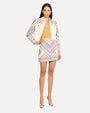 A dark-haired model wearing a yellow shirt under a white denim jacket with intricate, colorful embroidery on the shoulders and sleeves and a jean matching skirt. The model is wearing matching embroidered skirt with silver open-toed heels.