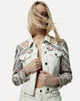 A waist up view of a blonde model wearing a white denim jacket with intricate, colorful embroidery on the shoulders and sleeves. The design is floral and geometric in blue, red and yellow colors. The jacket has metal buttons and two chest pockets with button flaps. 