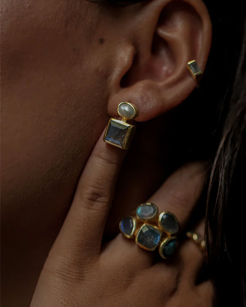 Close-up of an ear adorned with a gold earring featuring two stones—a small oval stone on top and a larger square stone below—and a gold ear cuff on the upper ear. A hand touching the earlobe is wearing multiple gold rings with blue stones.