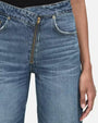 A zoomed-in view of a gold metal zipper on a pair of blue jeans. The zipper is positioned diagonally and is visible through the crossed waistband.