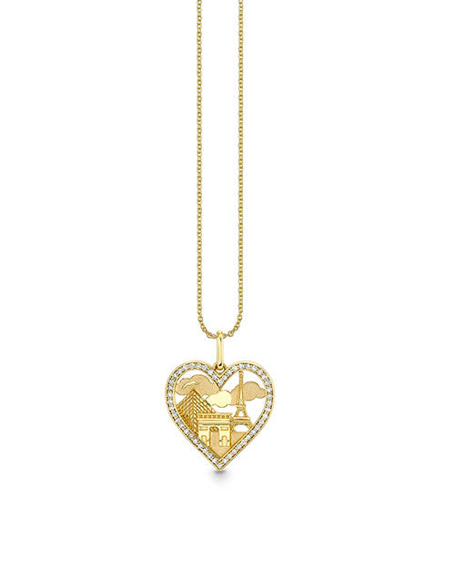 A gold heart-shaped pendant with intricate cut-out details hangs from a delicate gold chain. The pendant features a textured surface with various landmarks from France like the Eiffel tower and the Louvre.