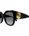 Close-up of black, oversized cat-eye sunglasses with grey-tinted lenses and a thick frame. On the temples, there is a bold circular gold emblem.