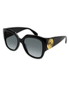 A pair of black, oversized cat-eye sunglasses with grey-tinted lenses and a thick frame. On the temples, there is a bold circular gold emblem.