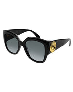A pair of black, oversized cat-eye sunglasses with grey-tinted lenses and a thick frame. On the temples, there is a bold circular gold emblem.