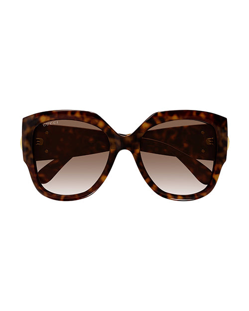 Front view of tortoiseshell colored, oversized cat-eye sunglasses with brown lenses and a thick frame. On the temples, there is a bold circular gold emblem.