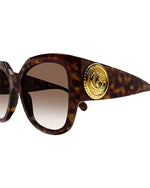 Close-up of tortoiseshell colored, oversized cat-eye sunglasses with brown lenses and a thick frame. On the temples, there is a bold circular gold emblem.
