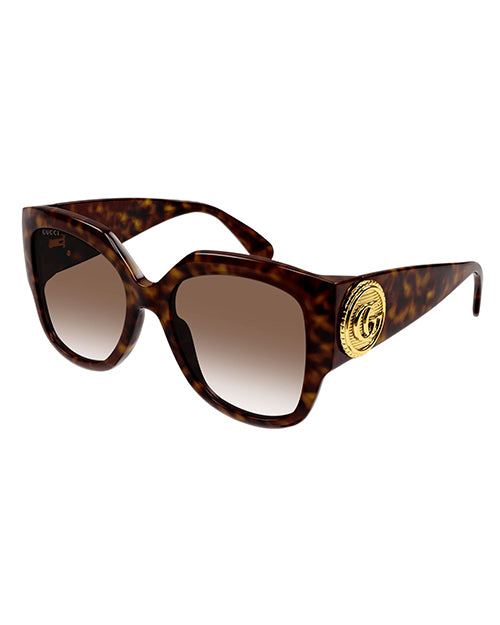 A pair of tortoiseshell colored, oversized cat-eye sunglasses with brown lenses and a thick frame. On the temples, there is a bold circular gold emblem.