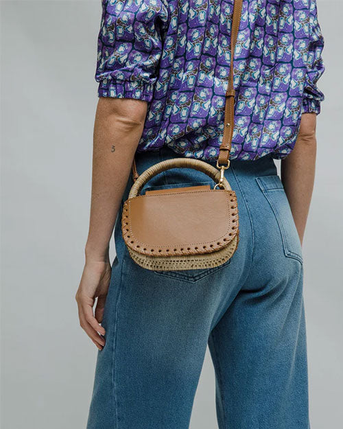 Person wearing a purple patterned shirt and blue jeans, carrying a small tan leather handbag with a rounded handle and decorative stitching along the edges.