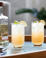 Two frosted glasses filled with a light orange cocktail are garnished with lemon and lime wedges on a table. A decorative bottle sits nearby.