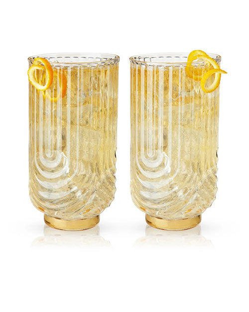 Two elegant, clear tumblers with vertical ridges, filled with a light beverage and ice, each garnished with a spiral of lemon peel. Sophisticated and refreshing.