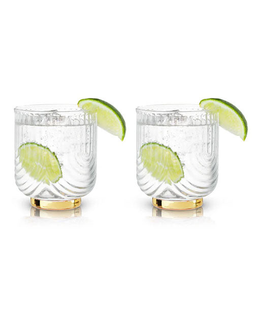 Two elegant glasses with striped details, each filled with a clear beverage and garnished with a lime wedge. Reflective gold bases add a touch of luxury.