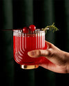 A hand holds a textured glass filled with a vibrant red cocktail, adorned with cranberries and a sprig of rosemary. The background is dark green, adding elegance.