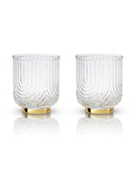 Two clear, textured glasses with vertical grooves and gold bases sit side by side on a reflective surface. The glasses have an elegant and luxurious feel.