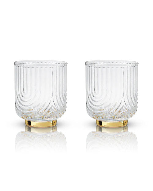 Two clear, textured glasses with vertical grooves and gold bases sit side by side on a reflective surface. The glasses have an elegant and luxurious feel.