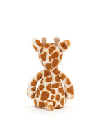 A back view of a plush toy giraffe sitting upright. The giraffe has brown spots on a cream-colored body and small horns.