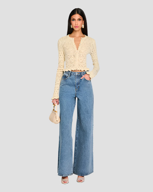 A person wearing a cream, long-sleeved, crochet cardigan with intricate patterns and scalloped edges along the hem, cuffs, and neckline. The cardigan features a variety of crochet patterns, including floral motifs and geometric shapes.   The cardigan is buttoned up in the front and paired with high-waisted blue jeans, a beige purse, and high heels.