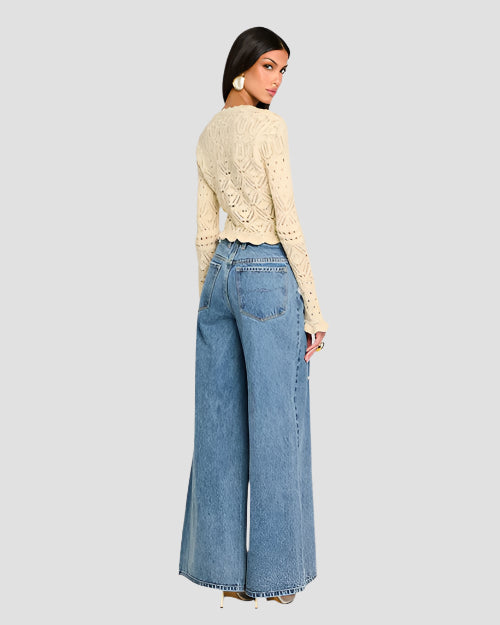 A backside view of a person wearing a cream, long-sleeved, crochet cardigan with intricate patterns and scalloped edges along the hem, cuffs, and neckline. The cardigan features a variety of crochet patterns, including floral motifs and geometric shapes. The cardigan is buttoned up in the front and paired with high-waisted blue jeans.