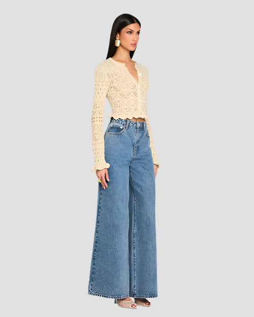 An angled side view of a person wearing a beige, long-sleeved, crochet cardigan with intricate patterns and scalloped edges along the hem, cuffs, and neckline. The cardigan features a variety of crochet patterns, including floral motifs and geometric shapes. The cardigan is buttoned up in the front and paired with high-waisted blue jeans.
