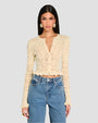 A person wearing a cream, long-sleeved, crochet cardigan with intricate patterns and scalloped edges along the hem, cuffs, and neckline. The cardigan features a variety of crochet patterns, including floral motifs and geometric shapes.   The cardigan is buttoned up in the front and paired with high-waisted blue jeans.