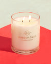 A lit scented candle from Glasshouse Fragrances is centered on a red surface with a beige background. The candle has two wicks, both of which are burning, and the wax appears to be white. The company’s logo and name are printed on the front of the glass container.